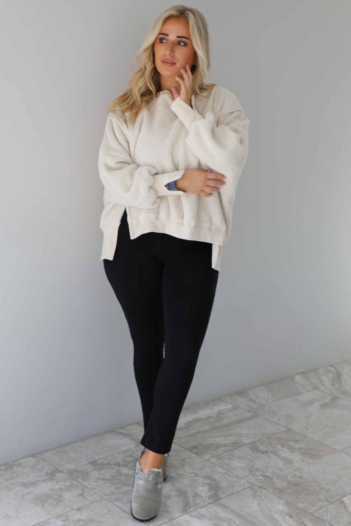 Woman wearing a sherpa sand oversized sweater with a fitted black jumpsuit underneath and grey slide on shoes. The photo is taken indoors against a white wall with white marble flooring. 