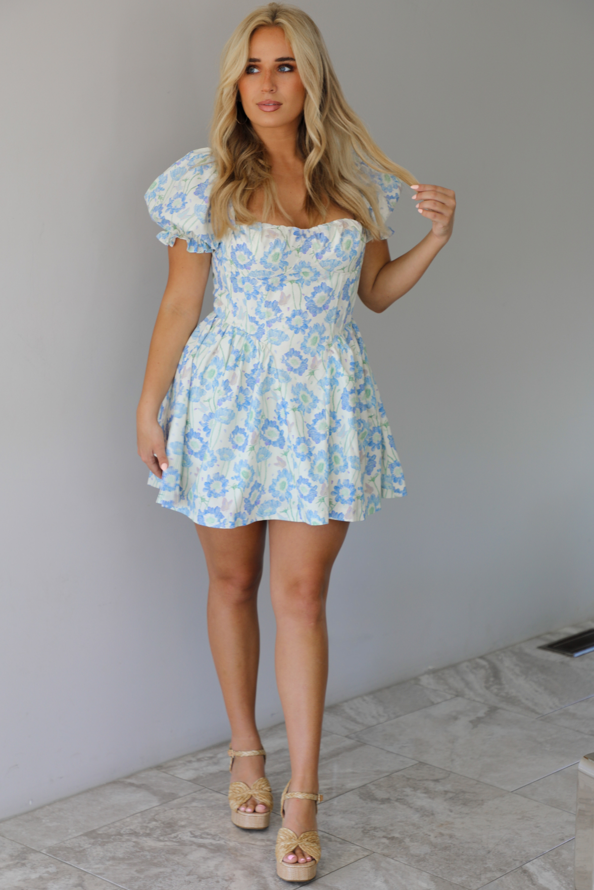 Woman wearing a white, blue, and green floral print mini dress that has short, puffed sleeves, a fitted corset bodice, and a flowy skirt indoors against a white wall with white marble flooring. 
