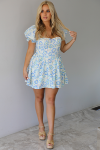 Woman wearing a white, blue, and green floral print mini dress that has short, puffed sleeves, a fitted corset bodice, and a flowy skirt indoors against a white wall with white marble flooring. 