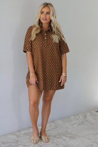 A woman modeling a brown short-sleeve mini dress with a cream polka dot pattern, standing indoors against a white wall with white marble flooring. 