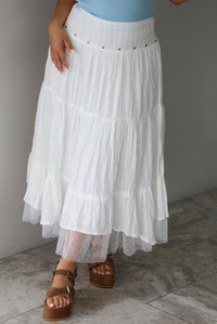 Woman wearing a white tiered mai length skirt with studded details at the waistline and sheer mesh detailing at the hem indoors against a white wall with white marble flooring. 