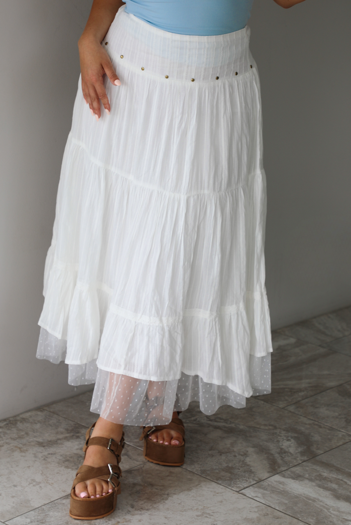 Woman wearing a white tiered mai length skirt with studded details at the waistline and sheer mesh detailing at the hem indoors against a white wall with white marble flooring. 