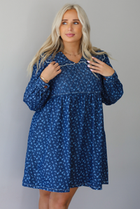 A woman wearing a dark denim mini dress with a white floral print pattern throughout, collared neckline, a quilted detail at the bodice, long sleeves, and a flowy skirt, standing indoors against a white wall.
