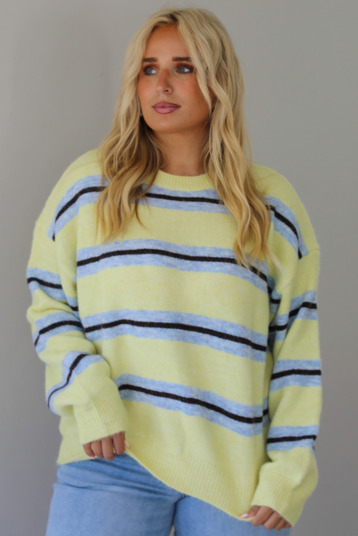 Woman wearing a pastel yellow, blue, and dark brown striped sweater, paired with light wash denim jeans that have a wide leg cut with a cuffed hem, standing indoors against a white wall.