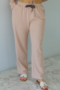 Woman wearing a tan two piece set with a hoodie top and sweatpants. The photo shows a closeup of the pants. The photo is taken indoors against a white wall.