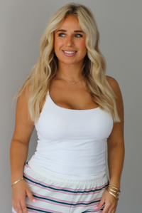 A woman modeling a pair of cream-white wide-leg pants with thin green and red horizontal stripes, paired with a white tank top, standing indoors against a white wall.