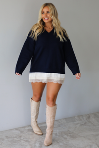 A woman wearing a navy sweater dress with a cream lace trim detail peaking out from underneath the dress giving it a layered look, paired with cream cowboy boots, standing indoors against a white wall with white marble flooring. 