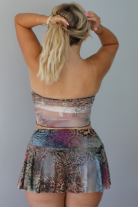 A blonde woman models a set with a tie-front corset-style bodice and a flowing multicolor abstract print skirt, standing indoors against a white wall. The photo shows the back of the set.
