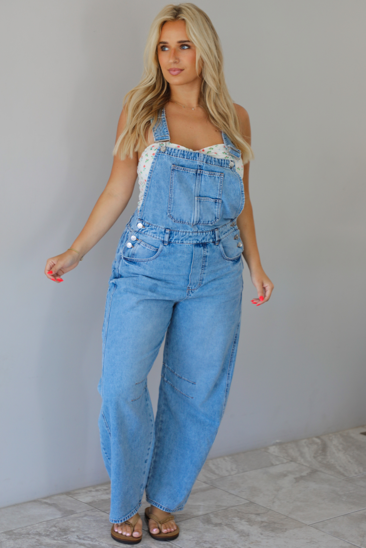Woman wearing a pair of denim overalls with a white floral strapless top underneath indoors against a white wall with white marble flooring. 