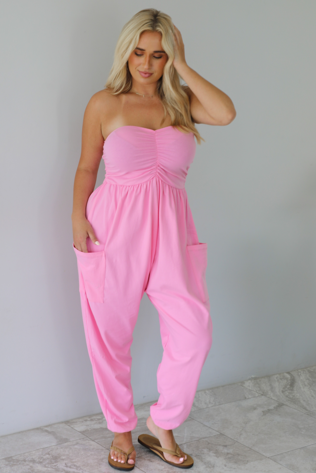 Woman wearing a pink strapless jumpsuit with a fitted bodice and slouchy pants indoors against a white wall with white marble flooring. 