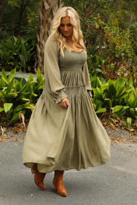 Woman model wearing an olive colored maxi length dress that has a smocked bodice, long sleeves that cinch with elastic at the wrists, and a flowy skirt. The photo is taken outdoors with greenery in the back.
