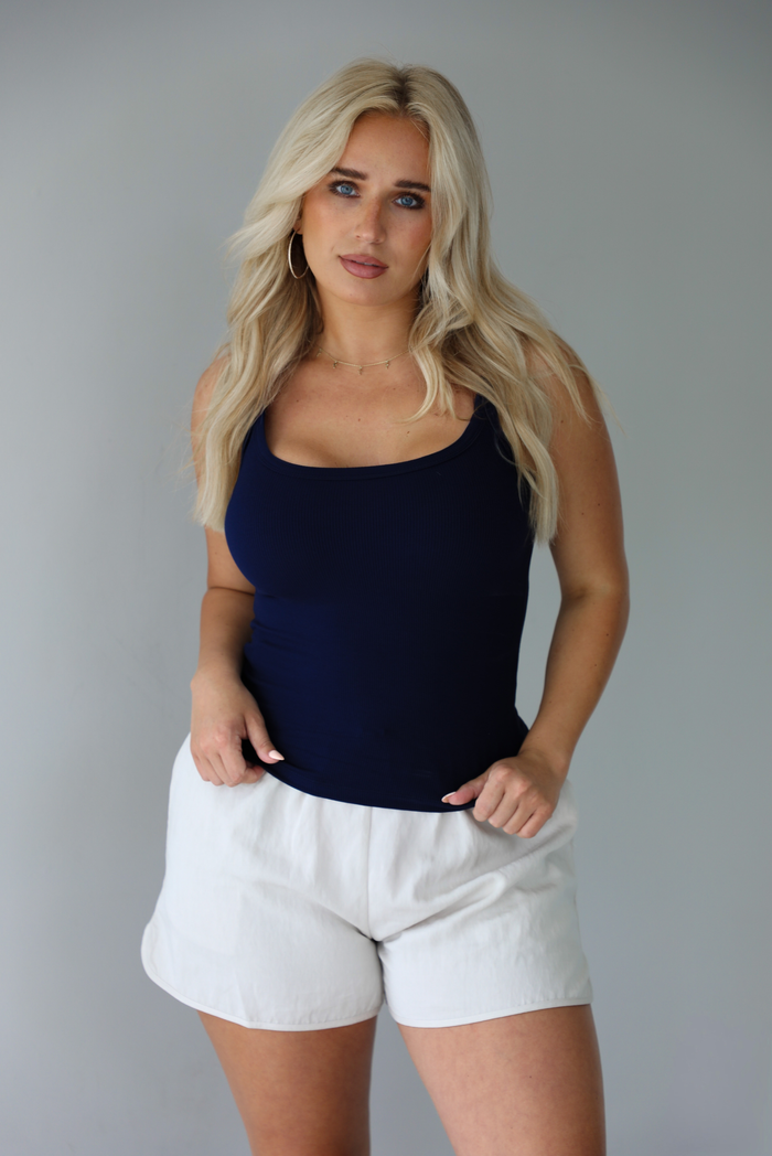 Woman wearing a navy ribbed tank  untucked with stone colored shorts indoors against a white wall. 