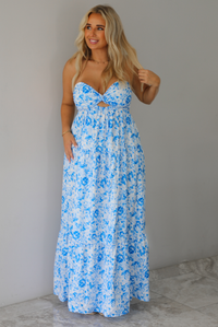 A woman modeling a strapless white maxi dress with a blue floral print and a sweetheart neckline with a small keyhole cutout, standing indoors against a white wall with white marble flooring. 
