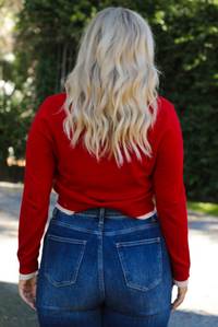 Woman model wearing a red cardigan with sand trim details and black button closures in the front with dark wash denim. The photo shows a close up of the back of the cardigan. The photo is taken outdoors on gravel with greenery in the back. 