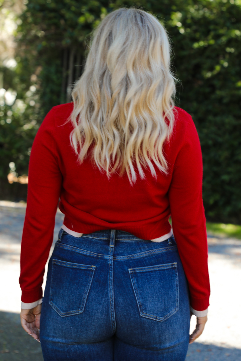 Woman model wearing a red cardigan with sand trim details and black button closures in the front with dark wash denim. The photo shows a close up of the back of the cardigan. The photo is taken outdoors on gravel with greenery in the back. 