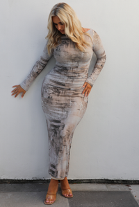 Woman model wearing a multi-colored and multi-patterned maxi length dress that is fitted with long sleeves. The photo is taken outdoors against a white wall. 