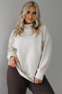 Woman wearing a beige high-neck, oversized sweaters and high waisted brown pants. The photo is taken indoors against a white wall with white marbled flooring. The photo shows a closeup of the oversized sweater untucked.