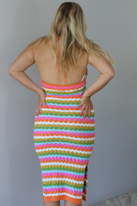 Woman wearing a bright, multi-colored striped midi dress with a halter neckline and a fitted design indoors against a white wall. The photo shows a closeup of the back, which is open, and has a halter tie at the top. 