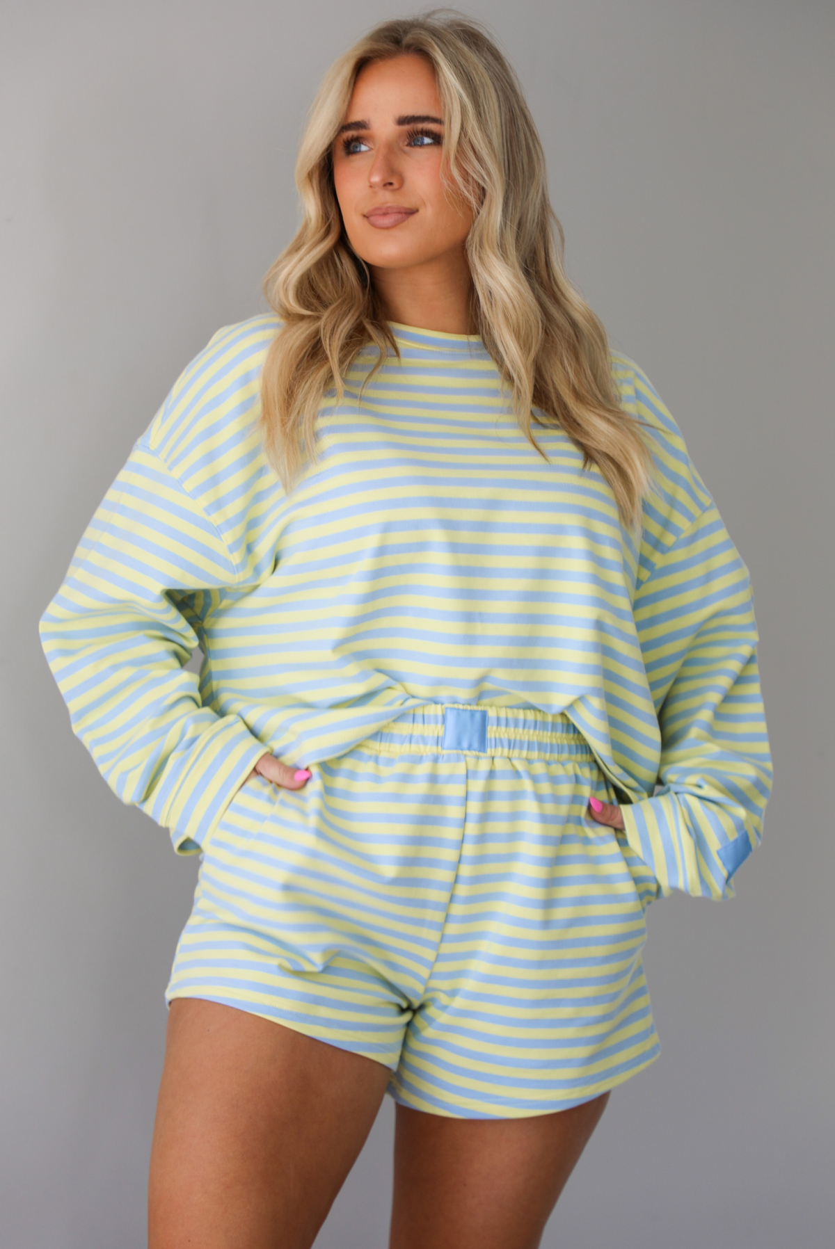 Woman wearing a blue and pale yellow striped two piece set featuring an oversized sweatshirt and shorts. The photo is taken indoors against a white wall and shows a closeup of the set, which is tucked into the front of the shorts. 