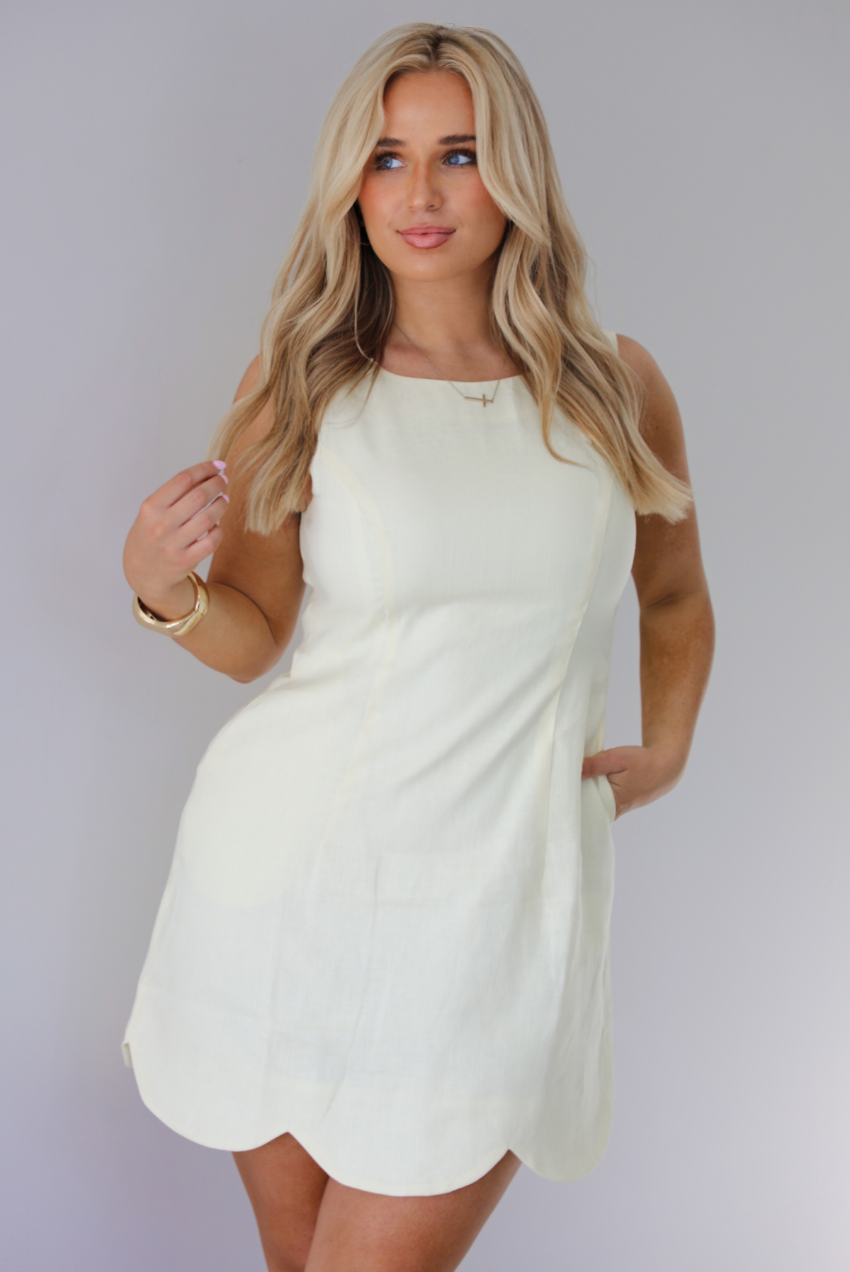 Woman wearing a pale yellow mini dress featuring a high neckline, sleeveless design, and a large scalloped hem. The photo is taken indoors against a white wall.