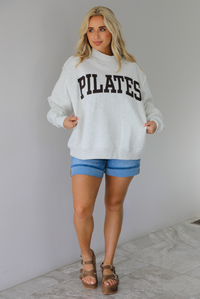 Woman wearing a heather grey oversized sweatshirt with the word 'PILATES' written in collegiate font on the front with medium wash denim shorts indoors against a white wall with white marble flooring. 