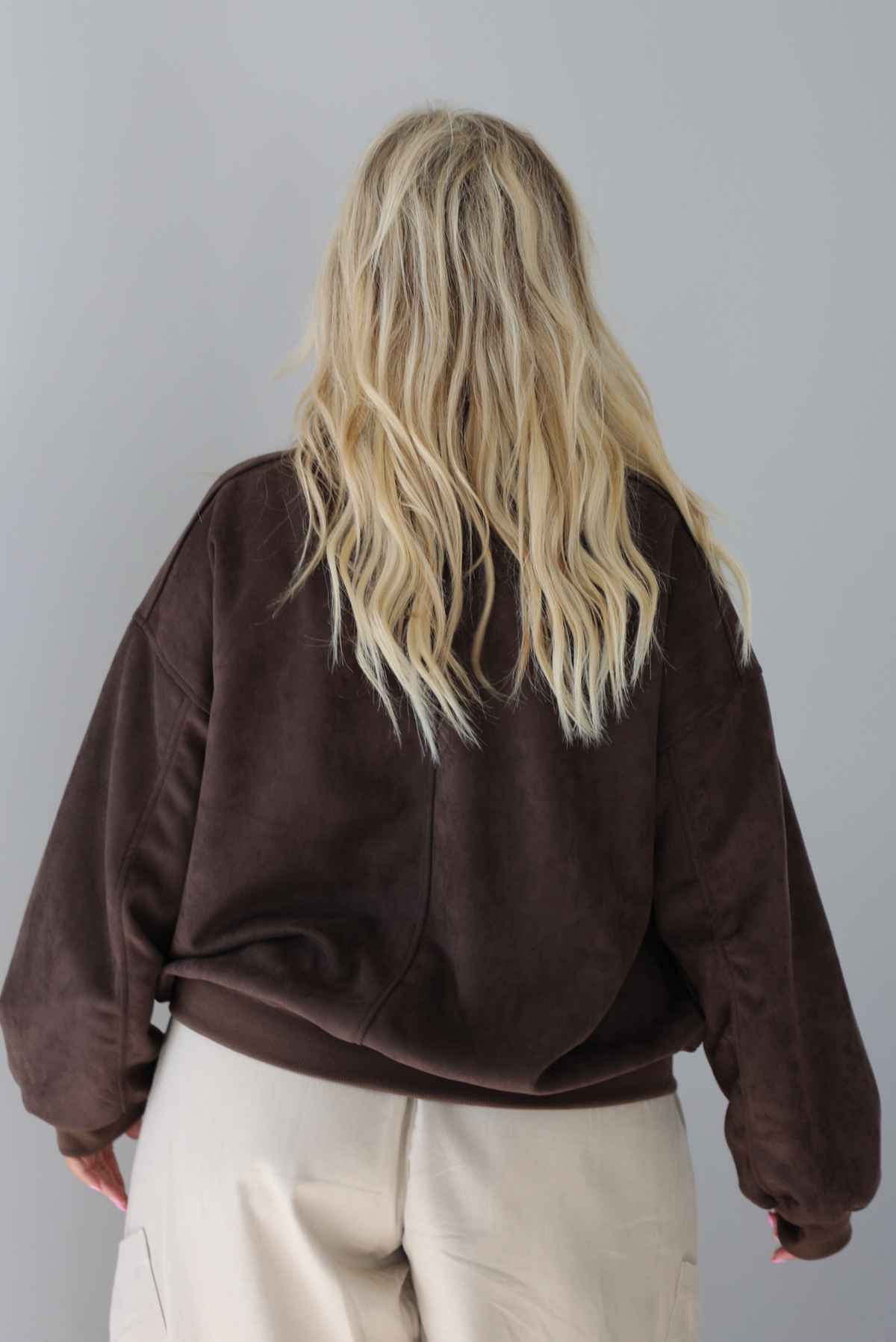 A woman shown from behind wearing a brown suede jacket with a white tank and sand colored baggy pants, standing indoors against a white wall. The photo shows a closeup of the back of the jacket. 