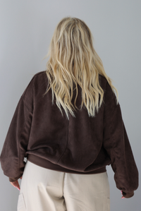 A woman shown from behind wearing a brown suede jacket with a white tank and sand colored baggy pants, standing indoors against a white wall. The photo shows a closeup of the back of the jacket. 