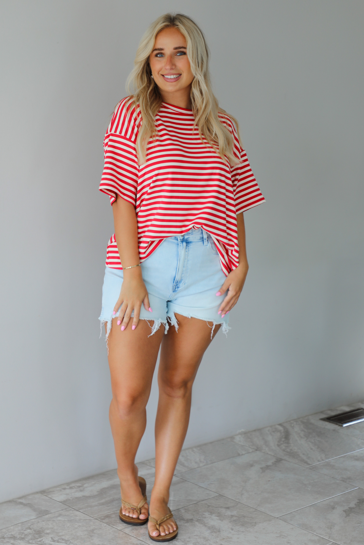 A woman modeling a red and white horizontally striped short-sleeve t-shirt with light blue denim shorts and brown sandals, standing indoors against a white wall with white marble flooring. 