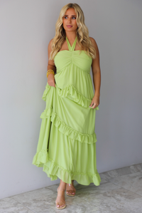 A woman modeling a lime green halter-neck maxi dress with tiered ruffle details, standing indoors against a white wall with white marble flooring. 