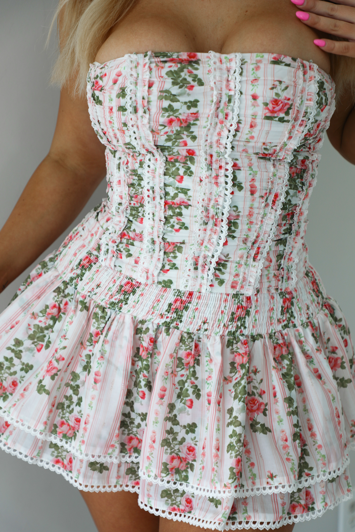 Woman wearing a white and pink floral two piece set featuring a strapless corset style top and an a ruffled skirt. The photo is taken indoors against a white wall.