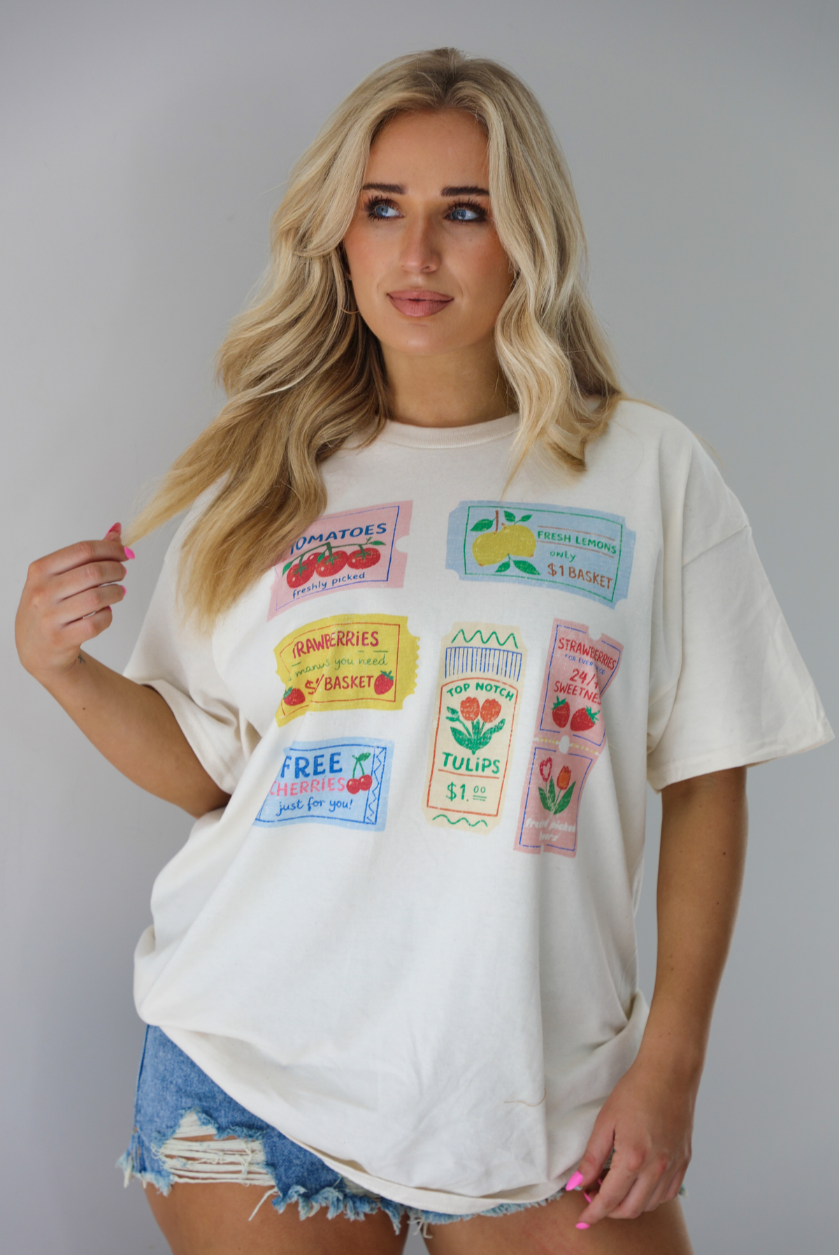 Woman wearing a white relaxed fit graphic tee that has various tickets on it with different fruits and flowers on them. The tee is untucked to show the full length and paired with distressed denim shorts. The photo is taken indoors against a white wall.