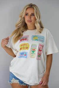 Woman wearing a white relaxed fit graphic tee that has various tickets on it with different fruits and flowers on them. The tee is untucked to show the full length and paired with distressed denim shorts. The photo is taken indoors against a white wall.