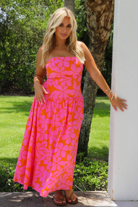 A woman wearing a bright pink and orange floral strapless maxi dress, standing outdoors next to a white pillar with a green lawn and palm tree in the background.
