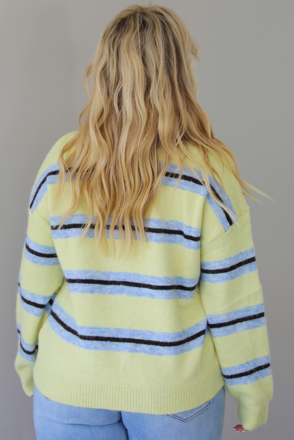 Woman shown from behind wearing a pastel yellow, blue, and dark brown striped sweater, paired with light wash denim jeans that have a wide leg cut with a cuffed hem, standing indoors against a white wall.
