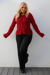 Woman model wearing a burgundy knit cardigan with button closures in the front and black denim jeans. The photo is taken outdoors against a white wall. 