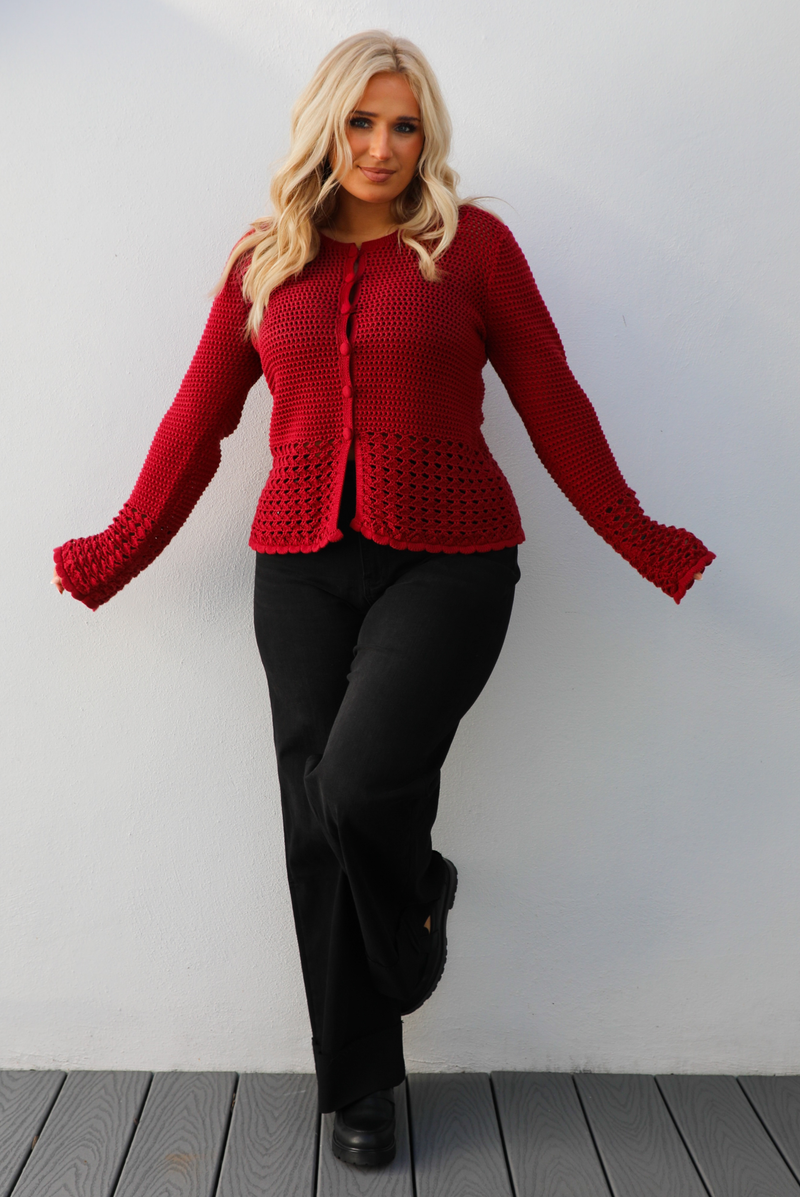 Woman model wearing a burgundy knit cardigan with button closures in the front and black denim jeans. The photo is taken outdoors against a white wall. 