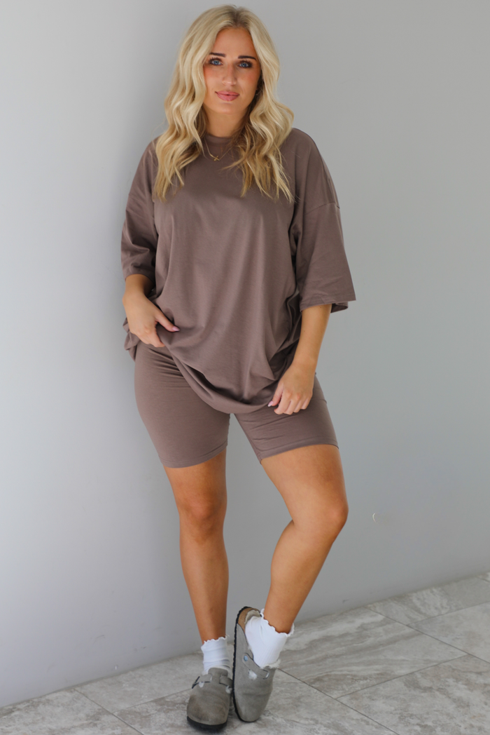 Woman wearing a two piece taupe set featuring an oversized t-shirt and fitted biker shorts. The set is paired with white ankle socks and slip on grey shoes. The photo is taken indoors in front of a white wall with white marble flooring.
