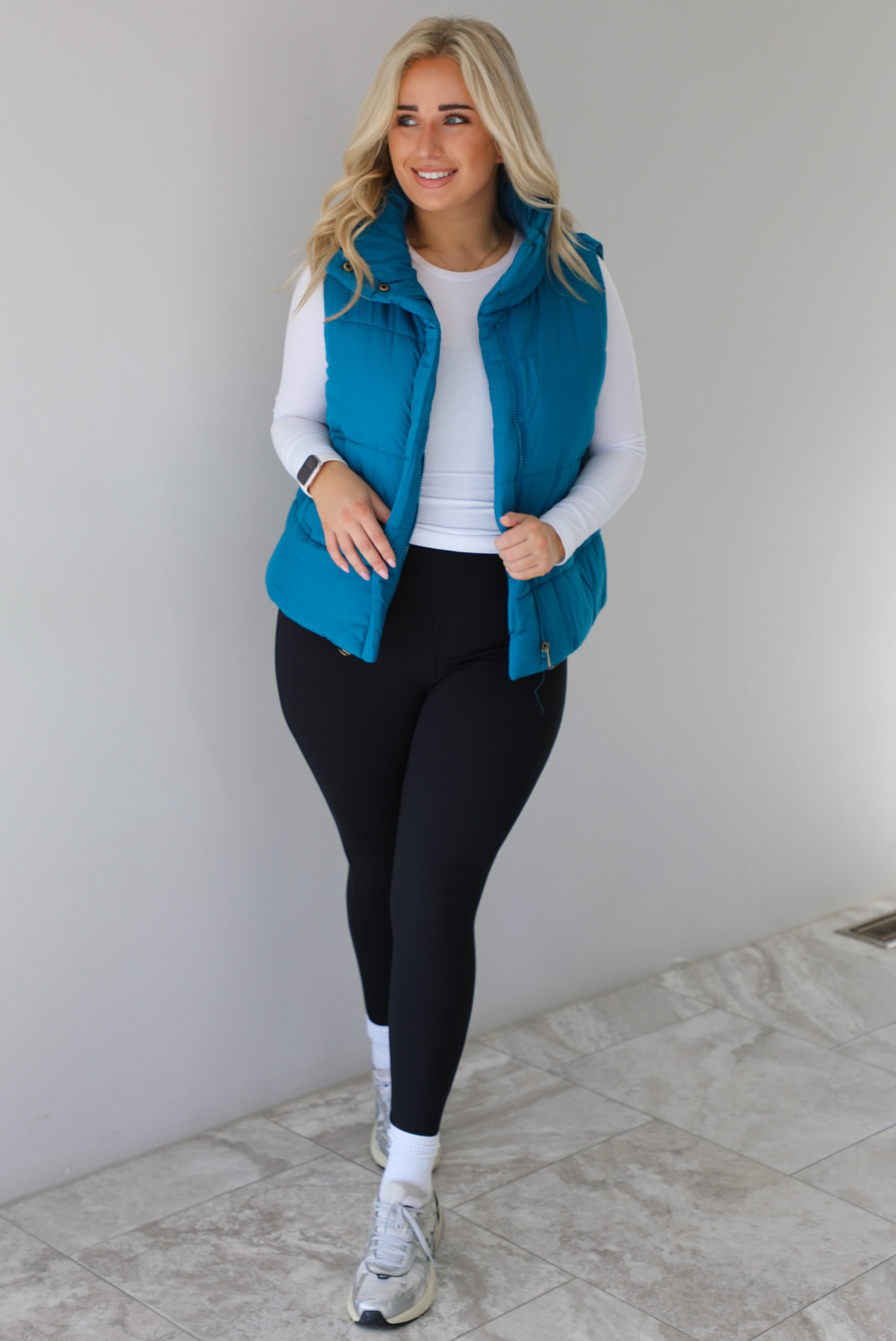 Woman wearing a teal puffer vest, white long-sleeve shirt, black leggings, and gray sneakers with white ankle socks indoors against a white wall with white marble flooring.