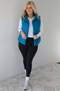 Woman wearing a teal puffer vest, white long-sleeve shirt, black leggings, and gray sneakers with white ankle socks indoors against a white wall with white marble flooring.