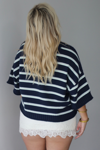 A woman shown from behind wearing a navy and multi-colored striped design knit top paired with a fitted cream, lace trim mini skirt, standing indoors against a white wall.