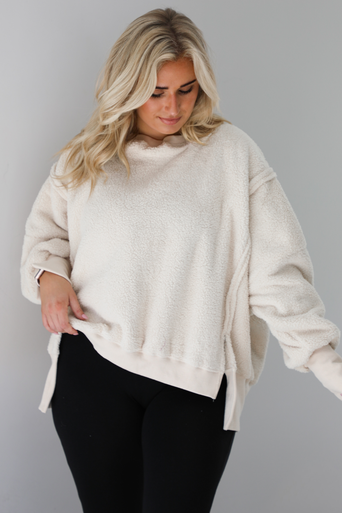 Woman wearing a sherpa sand oversized sweater with a fitted black jumpsuit underneath. The photo shows a closeup of the sweater and the raw seam details. The photo is taken indoors against a white wall with white marble flooring. 