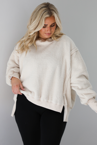 Woman wearing a sherpa sand oversized sweater with a fitted black jumpsuit underneath. The photo shows a closeup of the sweater and the raw seam details. The photo is taken indoors against a white wall with white marble flooring. 