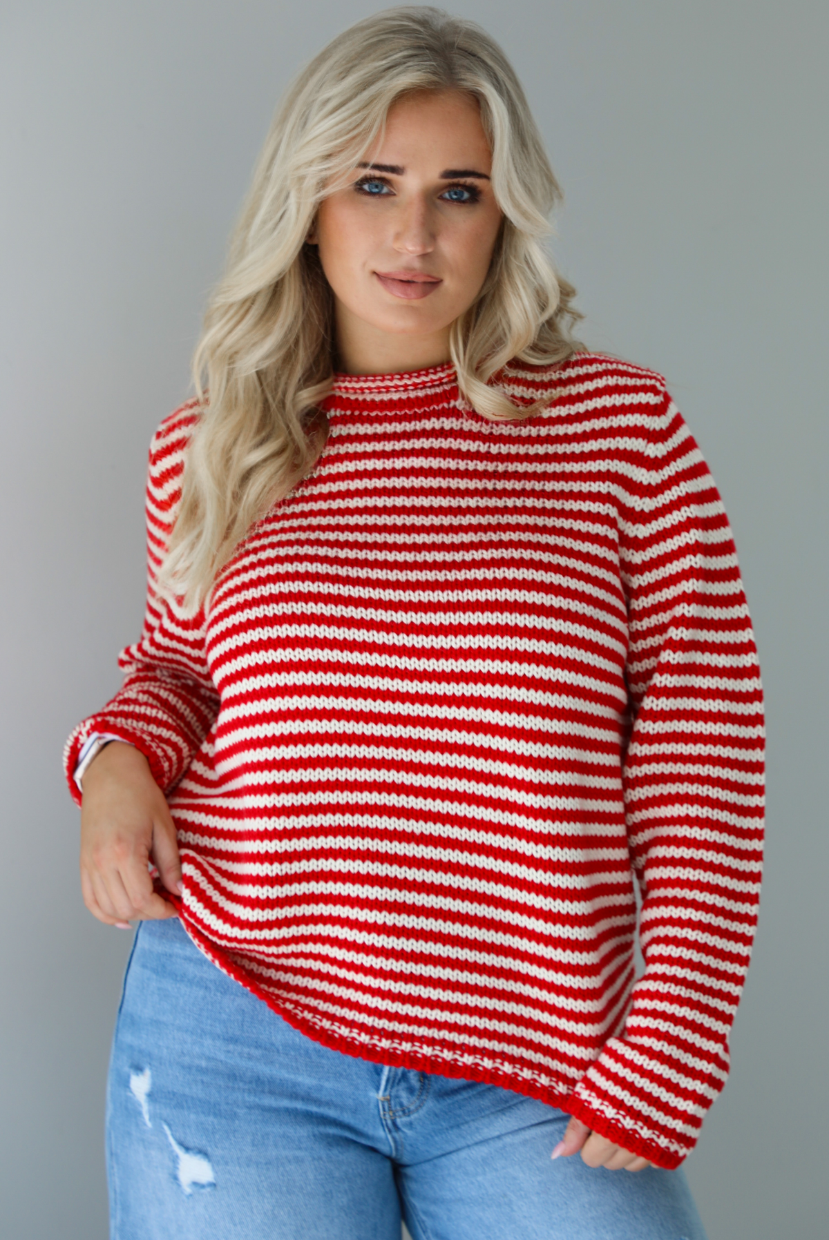 Woman wearing a red and white striped sweater with high waisted medium wash flared hem denim. The photo shows a closeup of the sweater untucked.  The photo is taken in front of a white wall.