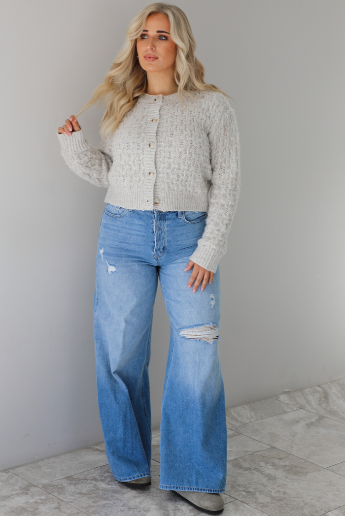 Woman wearing a light grey cardigan buttoned up with high waisted flared hem medium wash denim. The photo is taken indoors against a white wall with white marble flooring.