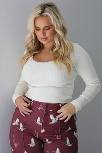 A woman wearing a cream colored knit long sleeved top paired with high waisted maroon jeans with a repeating mallard design, standing indoors against a white wall.
