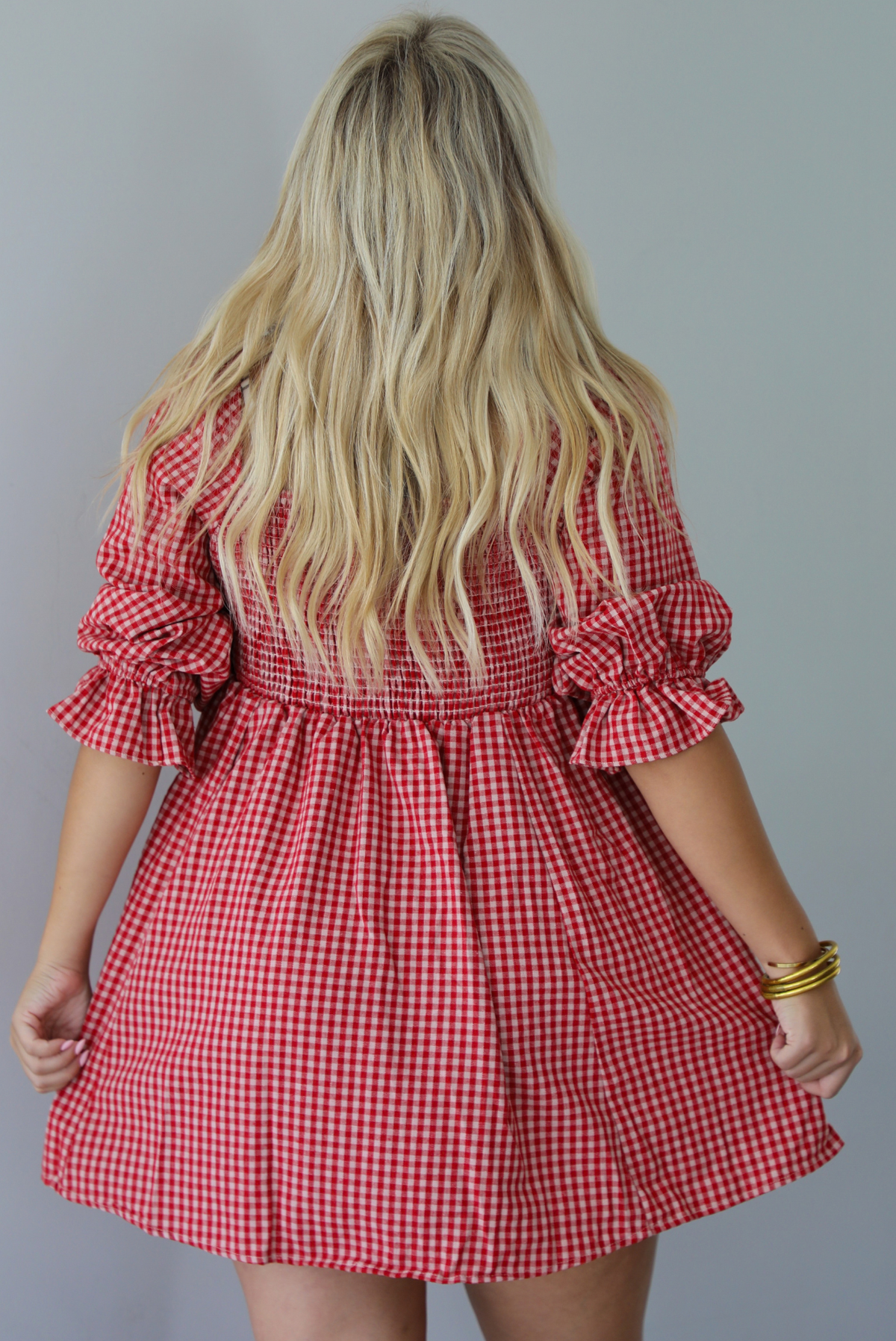 A woman shown from behind wearing a red and white gingham mini dress with puff, elbow length sleeves, standing indoors against a white wall.