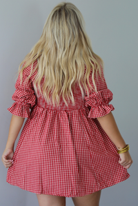 A woman shown from behind wearing a red and white gingham mini dress with puff, elbow length sleeves, standing indoors against a white wall.