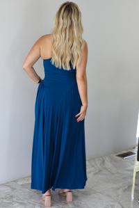 Woman wearing a blue one strap maxi dress indoors against a white wall with white marble flooring. The photo shows the back of the dress. 