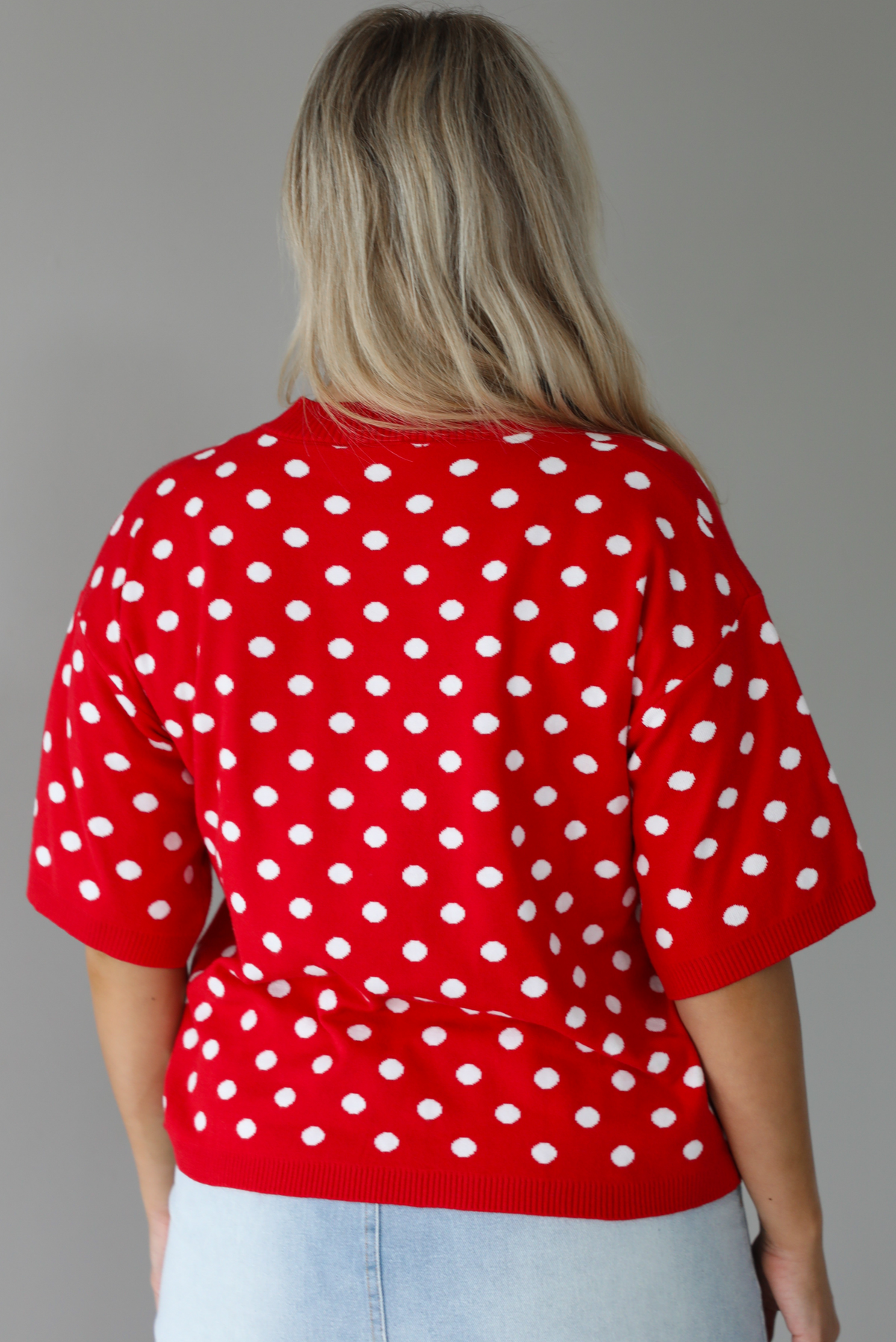 Woman wearing a red knit top with a white polka dot print pattern throughout with a high waisted, light-wash denim skort. The photo is taken indoors against a white wall and shows the back of the top. 