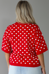 Woman wearing a red knit top with a white polka dot print pattern throughout with a high waisted, light-wash denim skort. The photo is taken indoors against a white wall and shows the back of the top. 